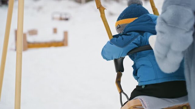 White mom pushes son on swing, blue jacket and pompom hat, closeup back view, protective hand guiding rope, wooden frame and snow underfoot, serene winter light, tender family bonding, calm