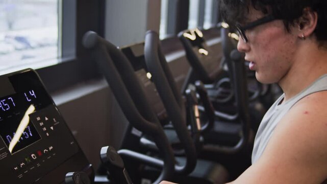 High angle shot of young Asian man using smartwatch to check heart beat while doing cardio on elliptical trainer