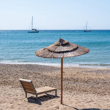 Straw umbrella and wooden lounger on the sand of Komi Beach, Chios Island, Greece. Summer sunny day, sailboats on sea.