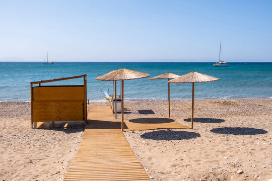 Sunny Mediterranean beach with straw umbrellas and sailboats on calm blue sea. Chios island, Greece