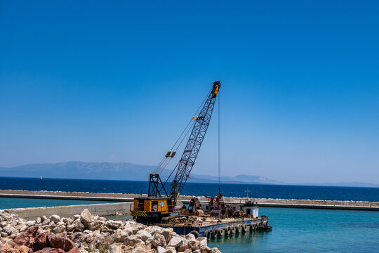Floating crane barge working near rocky pier in turquoise Greek island harbor under blue summer sky