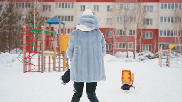 Playful snowy playground caucasian mom twirls son, colorful slide and climbing frame in background, legs kick and laughter, bundled snowsuits and hats, apartment facades behind trees, energetic