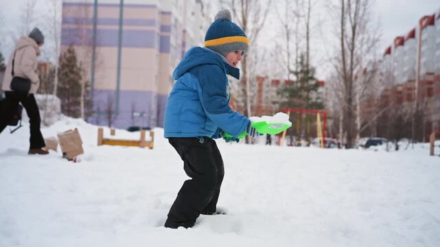 White son with green shovel digging, urban playground near apartment buildings mom watching from distance bundled in blue jacket and knit hat playful scooping and piling cold overcast daylight family