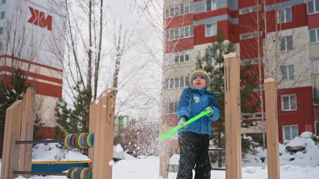 White son holding green shovel amid snow bundled in blue jacket and knit hat against apartment backdrop light flurries steady gaze exploring wooden play structure urban winter mood playful resilience