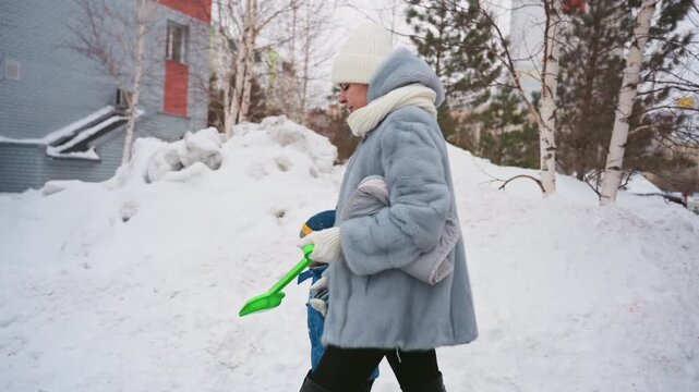 White mom and son walking snowy sidewalk, mom carrying bright green shovel and bundled child in blue jacket crisp winter day with snowbanks, birch trees and apartment facades cozy coat, knit hat,