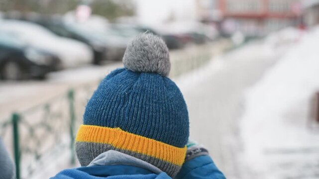 Closeup of white son knit hat and back, blue beanie with yellow stripe and grey pom, walking beside mother on snowy sidewalk, wool texture, cozy winter detail, playful childhood moment, urban stroll