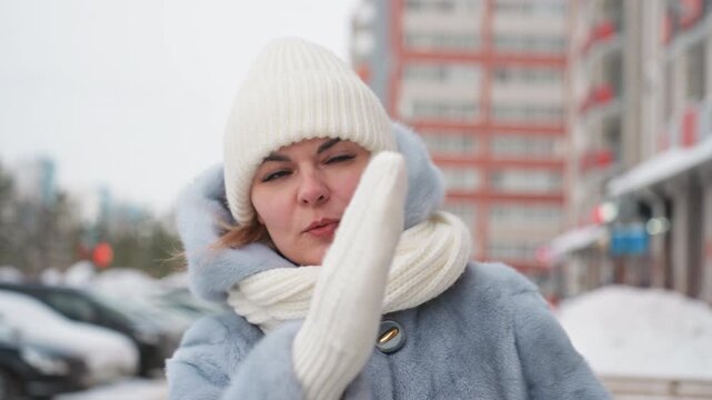 Caucasian mom calling son on snowy city sidewalk, wearing white knit hat, cream scarf and blue coat. Expressive hand gestures and concerned facial expression, visible breath in cold air. Urban