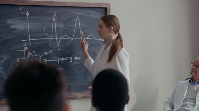 Medium shot of Caucasian female student standing at chalkboard while explaining anatomy theory to senior teacher and classmates in modern classroom