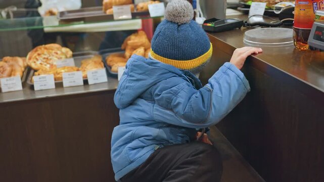 Caucasian son reaches bakery display with mom nearby, curious toddler in blue coat and yellowstriped beanie stretching to grasp counter edge and peer at pastries mother stands close and watchful,