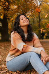 Obraz premium A young woman sits joyfully amongst falling autumn leaves in a park, with her eyes closed and a peaceful smile, radiating contentment.