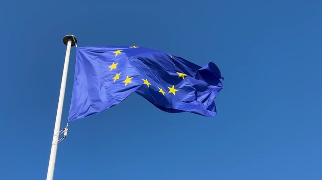 The European Flag on a large flagpole flying and flapping in the wind with a blue sky