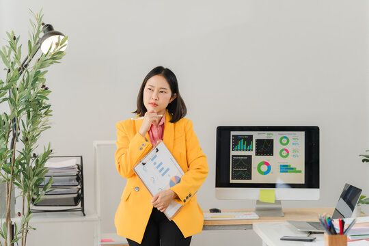 Introspective Moment: An office setting with an Asian businessperson contemplating strategy, holding documents, standing next to a computer displaying data analysis.