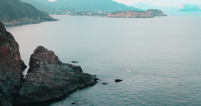 Aerial view of two kayakers in calm blue ocean. Rugged rocky cliffs and distant city skyline. Serene coastal landscape. High-angle travel photography.
