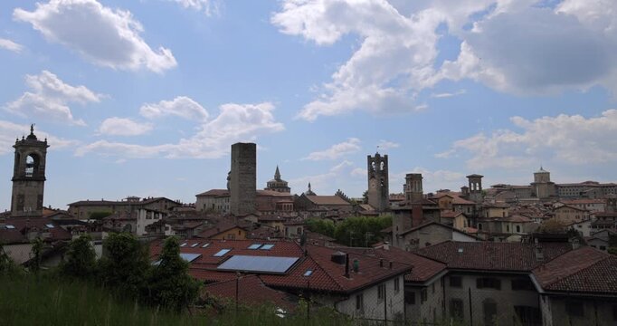 Static panorama of Bergamo Alta rooftops and landmark towers under summer clouds, locked off view holds layered medieval skyline with terracotta roofs and green hills in the distance