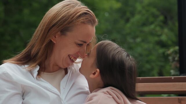 Tender mom daughter nose touch bench, gentle laughter and eye contact in park, warm closeup capturing playful affection and soft smiles, natural greenery background and casual white shirt detail