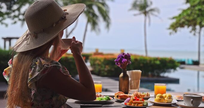 Woman wearing straw hat drinking coffee at tropical resort breakfast table, ocean and palm tree view, luxury vacation, relaxing morning lifestyle, travel tourism.