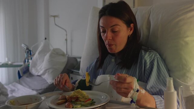 Close view of female patient eating in hospital bed with intravenous catheter visible, lifting fork with food, expression neutral and tired suggesting recovery and healing