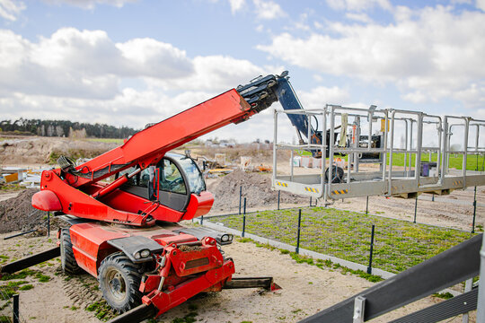 Red telescopic handler lifting a platform above green crops on a construction site with blue sky and clouds in the background