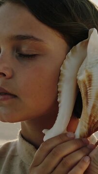 Vertical video: Raising shell girl long-sleeve top pressing conch to right ear hearing sea at shore