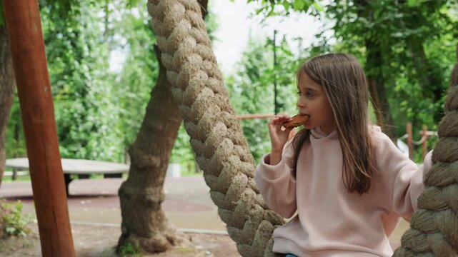 Daughter eating cookie on rope swing in park, braided hair and cozy sweater, crumbs on fingers, expressive gaze and content expression while savoring treat during peaceful outdoor moment