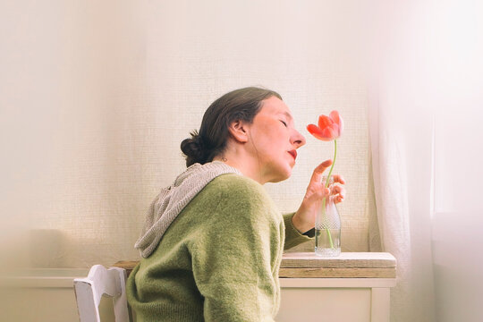 Woman sitting in front of a window in a meditative and romantic pose with a red tulip in her vase on a wooden shelf in the background VII