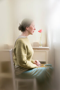 Woman sitting in front of a window in a meditative and romantic pose with a red tulip in her vase on a wooden shelf in the background VIII