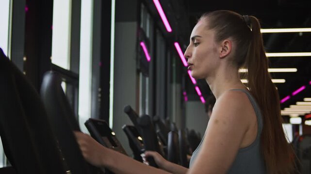 Side view shot of young Caucasian woman breathing heavily while using elliptical trainer after workout in gym
