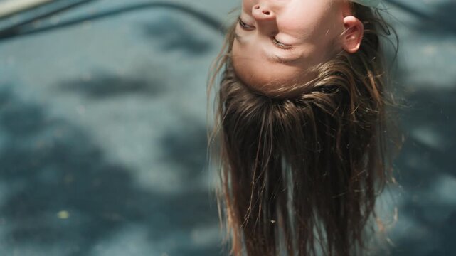 White daughter hanging upside down on playground, close portrait of braided hair swinging through sunlit shadows, candid joyful expression, summer outdoor play and carefree childhood moment.