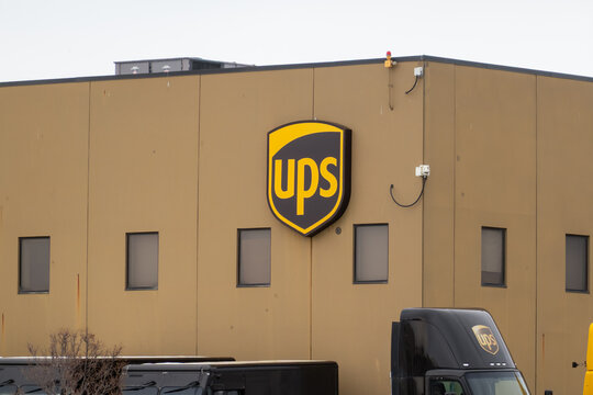Calgary, Alberta, Canada. April 7, 2026. UPS logo shield on a brown distribution center building with delivery trucks parked in the foreground.