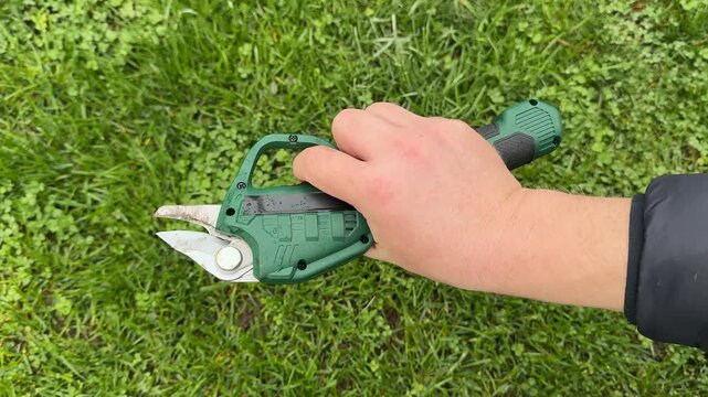 Closeup of hand holding electric pruning shears over green lawn, outdoor gardening maintenance task, spring yard work tool for trimming plants