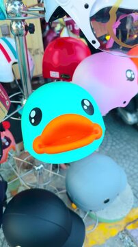 Cute blue duck face helmet displayed among various colorful motorcycle and scooter protection gear accessories, inviting riders to choose a fun and safe option for urban commuting