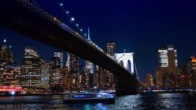 Modern riverboat passes under the Brooklyn Bridge at night. Dazzling skyline of Manhattan, New York City, US in the backdrop.