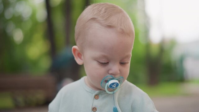 White toddler closeup with pacifier looking down in garden setting, knit sweater and soft bokeh background create serene mood, reflective parenting snapshot highlighting early emotion and lifestyle
