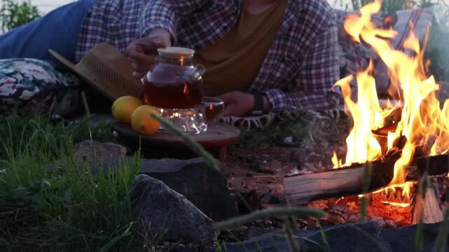 Pouring Tea from Glass Teapot by Campfire, Cozy Outdoor Evening. Person pouring hot tea from glass teapot near campfire. Cozy outdoor evening and relaxation concept