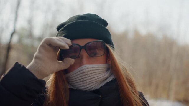Redhead caucasian woman adjusts sunglasses while scarf covers lower face. Beanie and backpack visible on winter forest trail. Gloved hand lifts glasses to reveal eyes in closeup. Cinematic backlight