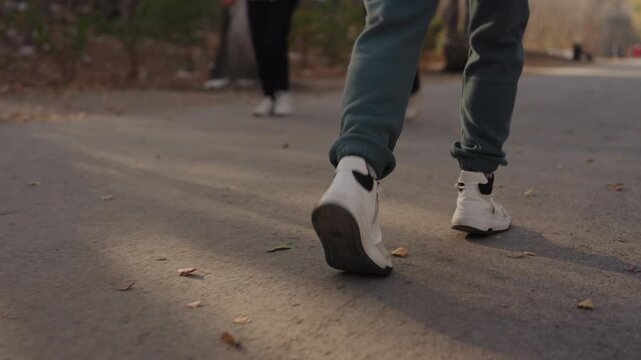 Closeup footsteps on park pavement. Pedestrian in green pants and white sneakers walking away, camera follows stride from rear, distant figure suggests approaching prowler or threat, warm autumn light