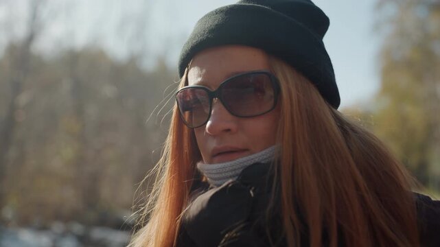 Woman in cold park. Redheaded woman wearing scarf sunglasses. Detailed shot of redheaded woman in scarf and sunglasses breathing out visible air in winter park with hints of surveillance