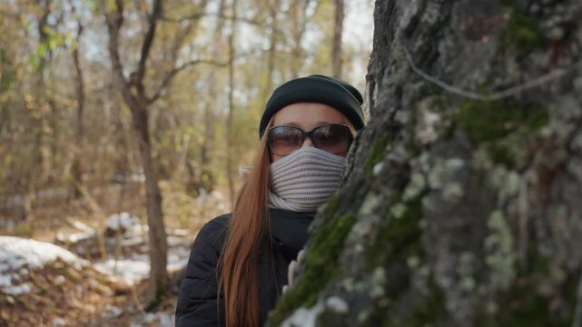 Caucasian redhead woman peeking from behind birch, masked and bundled in scarf and beanie, sunglasses hiding expression, mossy trunk closeup, snowdusted ground, lingering suspense suggesting stealthy