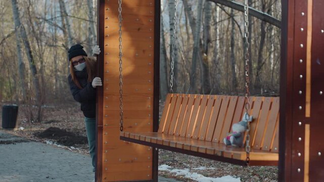 White female prowler peeks around wooden shelter then snatches plush rabbit from swing bench, covert approach through park trees, tense glance and hurried reach, backpacked passerby acting