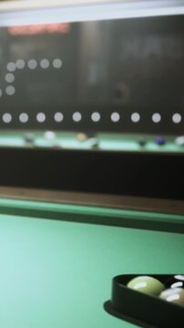 A green pool table sits in a dark room with several balls arranged in a triangle. Players are preparing for a friendly match while lights shine above the table.