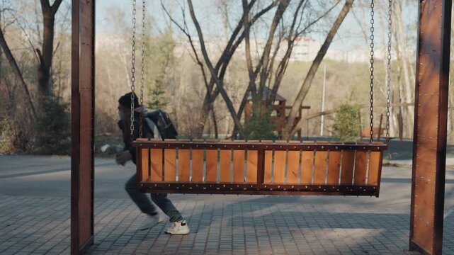 Park swing bench snatcher slips behind wooden seat, paved plaza with bare trees, hooded figure moves furtively along chains, quick glance over shoulder, silent getaway in late afternoon light,
