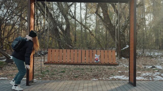 Sneaking along snowy park bench, backpacked person furtively approaches and snatches small object, cautious glances, cold late afternoon, leafstrewn trail and wooded backdrop create tense mood.
