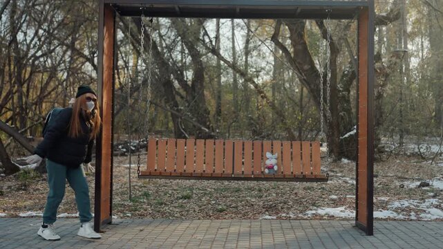 Suspicious person approaches swing bench, inspects unattended bag then snatches it and flees along paved park path autumn trees and scattered leaves create tense outdoor atmosphere as figure in beanie