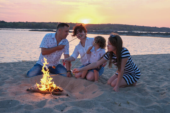 Happy family gathered around a beach bonfire at sunset, parents and kids roasting marshmallows, sharing laughs and creating warm summer memories by the lakefront