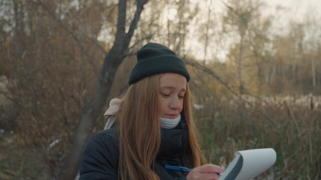 Redhead caucasian woman hides paper pad outdoors in marshland beanie and scarf visible flipping pages and peeking around trees pensive expression field researcher turned nature illustrator taking