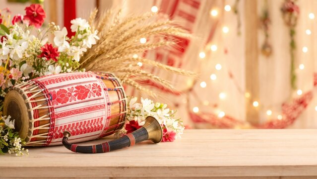 Festive Bihu background with traditional dhol drum, pepa horn, and wheat stalks on a wooden table. Warm bokeh lights and gamosa fabric in the background with large copy space for text or design.