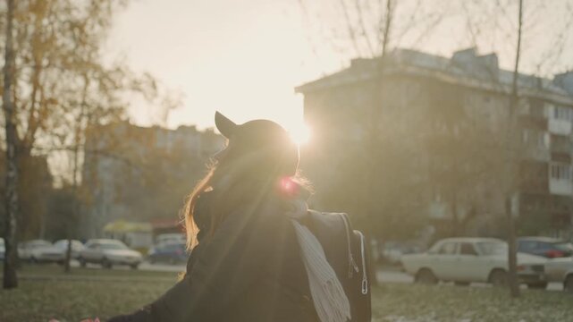 Caucasian redhead woman holding plush toy toward skyline at golden sunset, winter jacket and cap, backpack and gloves visible, sunbeam rim light creates warm halo, playful protective expression, soft