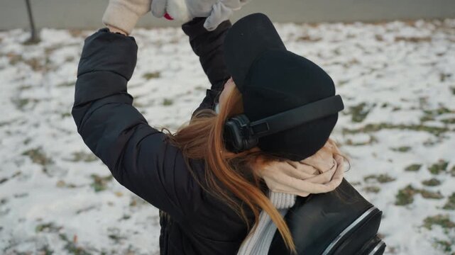 Caucasian woman celebrates reclaiming plush bunny in snowy park, arms raised in victory, joyful expression with headphones and scarf, backpack on shoulders, closeup on coat and jeans, walking