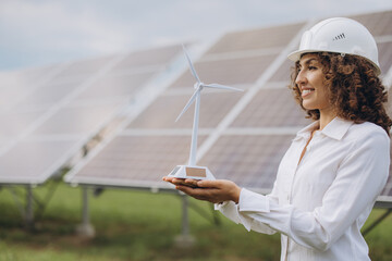 Young woman in hard hat presenting a wind turbine model, standing in front of solar panels,...