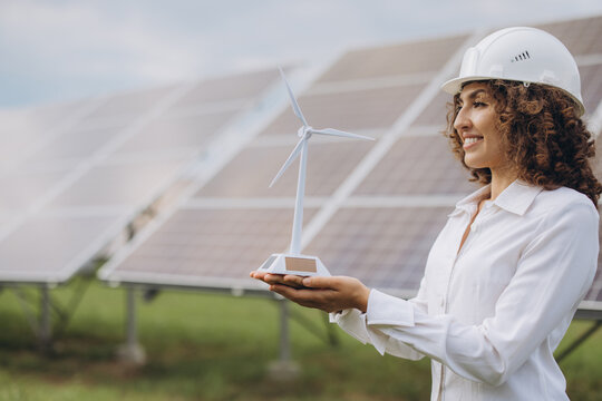 Young woman in hard hat presenting a wind turbine model, standing in front of solar panels, planning green technology